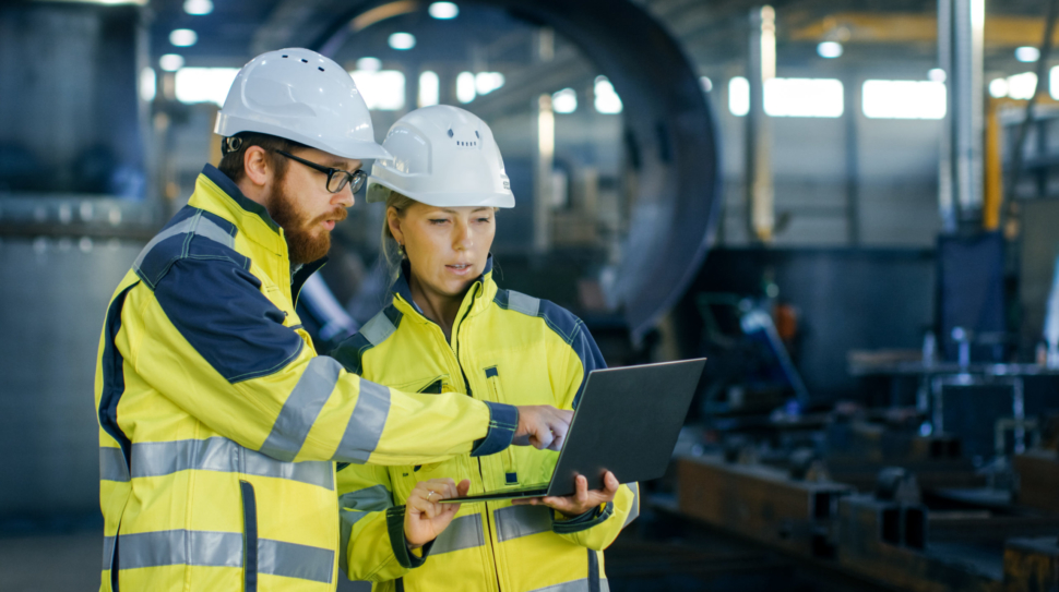 Male and Female Industrial Engineers in Hard Hats Discuss New Project while Using Laptop. They Make Showing Gestures.They Work in a Heavy Industry Manufacturing Factory.